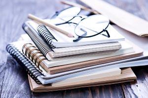 Una pila de cuadernos con un lápiz y un par de gafas encima, sentados en un escritorio de madera