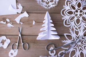Paper cut into the shapes of Christmas trees and snowflakes sitting on a wood table with two pairs of scissors