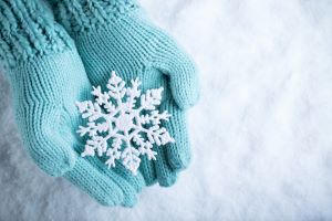 A pair of hands wearing light blue gloves holding an intricate snowflake decoration over a snow covered background